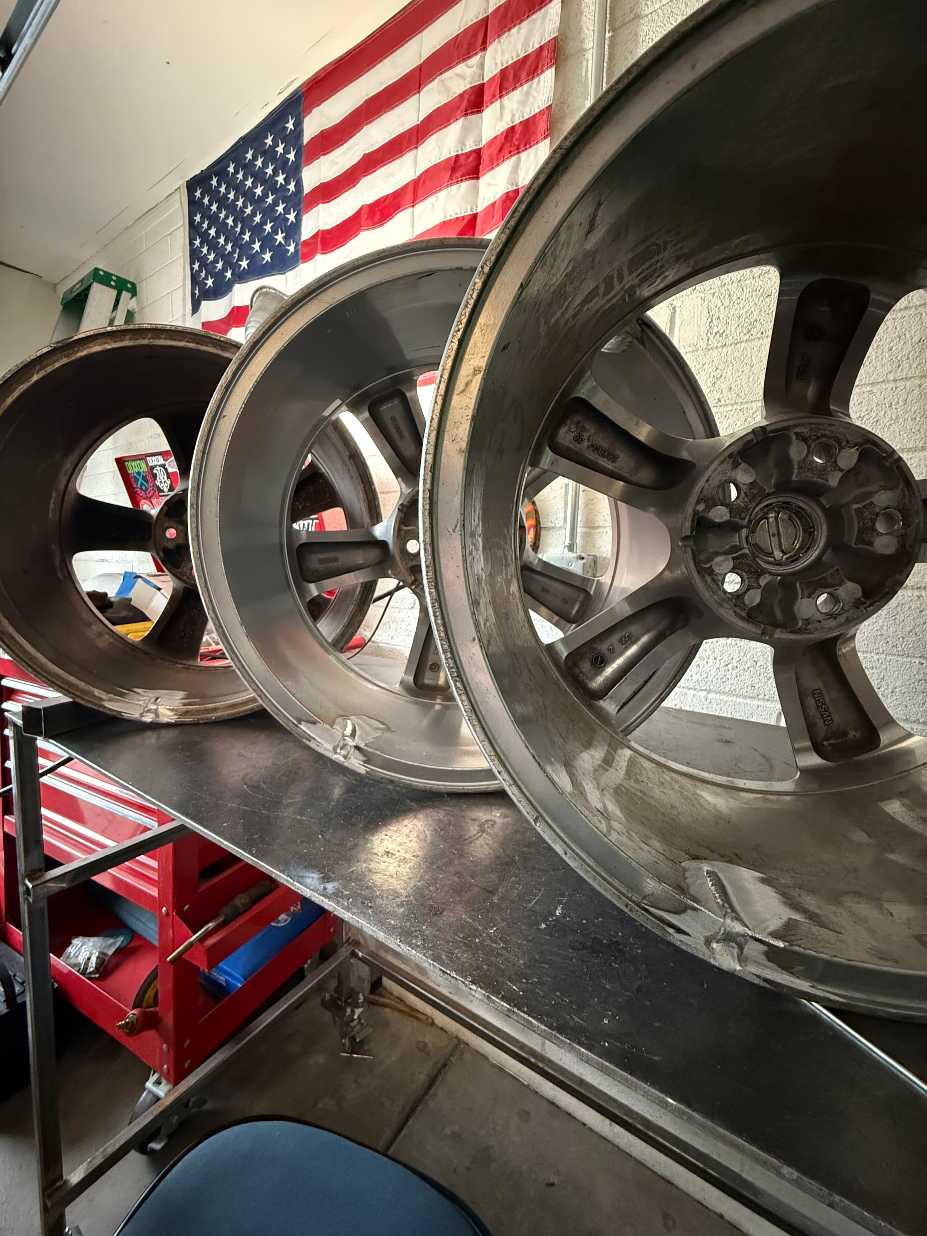 Three silver Nissan wheels on a metal workbench in a garage with an American flag.