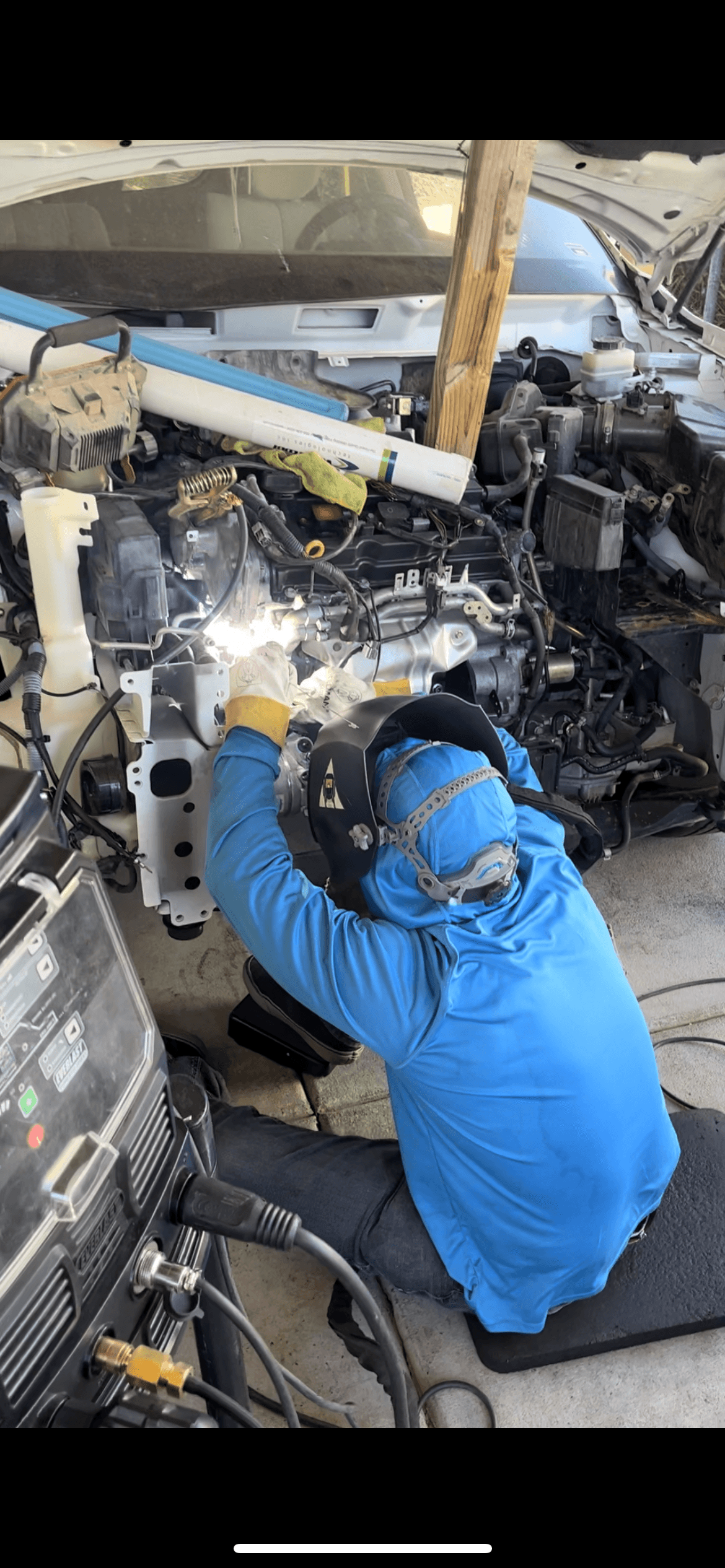 A welder in a blue shirt performs repairs inside a white car's engine compartment.